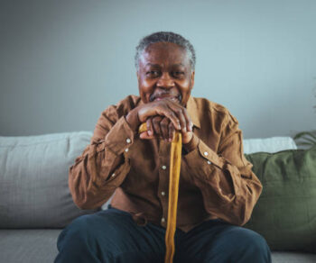 Portrait of happy senior man smiling at home while holding walking cane. Old man relaxing on sofa and looking at camera. Portrait of elderly man enjoying retirement.
