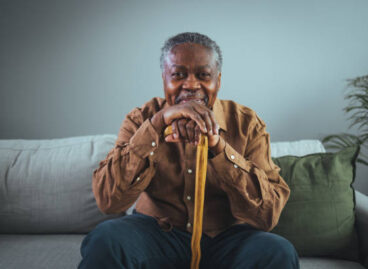 Portrait of happy senior man smiling at home while holding walking cane. Old man relaxing on sofa and looking at camera. Portrait of elderly man enjoying retirement.