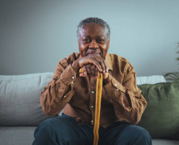 Portrait of happy senior man smiling at home while holding walking cane. Old man relaxing on sofa and looking at camera. Portrait of elderly man enjoying retirement.