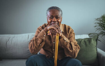 Portrait of happy senior man smiling at home while holding walking cane. Old man relaxing on sofa and looking at camera. Portrait of elderly man enjoying retirement.