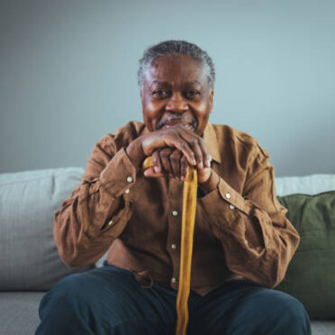 Portrait of happy senior man smiling at home while holding walking cane. Old man relaxing on sofa and looking at camera. Portrait of elderly man enjoying retirement.