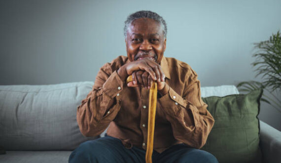 Portrait of happy senior man smiling at home while holding walking cane. Old man relaxing on sofa and looking at camera. Portrait of elderly man enjoying retirement.