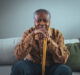 Portrait of happy senior man smiling at home while holding walking cane. Old man relaxing on sofa and looking at camera. Portrait of elderly man enjoying retirement.